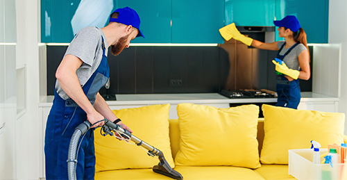 young man with vacuum cleaner washing with vapor yellow sofa beautiful woman wipes cuisine furniture